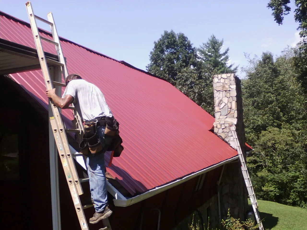 Roofer on a ladder beside a red metal roof and stone chimney — Soffit & Fascia Repair by Roofing Brothers in Cadiz